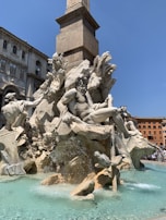 A detailed stone sculpture featuring mythological figures is set against a clear blue sky. The central figure is a muscular man reclining on rocks, surrounded by other sculpted figures and water flowing around them. The background includes an ancient, architecturally detailed building and a tall stone obelisk.