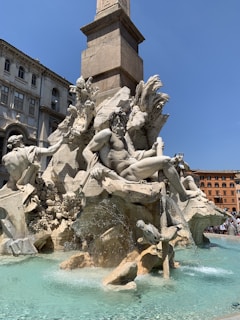A detailed stone sculpture featuring mythological figures is set against a clear blue sky. The central figure is a muscular man reclining on rocks, surrounded by other sculpted figures and water flowing around them. The background includes an ancient, architecturally detailed building and a tall stone obelisk.