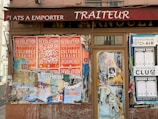 A storefront with a maroon-colored awning displaying the word 'TRAITEUR' and partially visible text 'PLATS A EMPORTER' above it. The facade is covered with a collage of overlapping posters advertising various events, with prominent orange posters promoting a 'Revolution' and 'Cultures Urbaines' festival. Other posters include mentions of 'Open Air' and 'Dehors Brut'. The posters are in different colors and are in various states of deterioration.