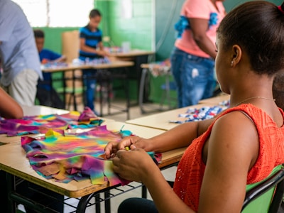 A group of people are engaged in crafting activities in a classroom setting. A woman in the foreground is focused on making something with colorful fabric pieces on a desk. Other individuals in the background are also occupied with crafting materials at their respective desks. The room has a vibrant atmosphere with green walls and natural light filtering through the windows.