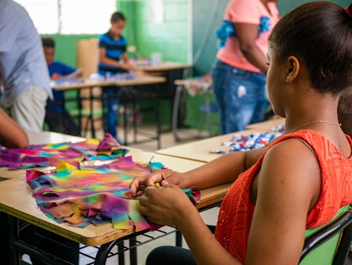 A group of people are engaged in crafting activities in a classroom setting. A woman in the foreground is focused on making something with colorful fabric pieces on a desk. Other individuals in the background are also occupied with crafting materials at their respective desks. The room has a vibrant atmosphere with green walls and natural light filtering through the windows.