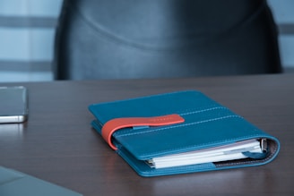 A teal notebook with an orange strap rests on a wooden table. The notebook is closed, and a smartphone is visible nearby. In the background, a black leather chair can be seen.