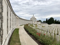 An expansive war memorial site with a long, curving wall of engraved names. A central domed structure stands prominently in the distance, surrounded by neatly arranged rows of white gravestones. The foreground features a path bordered by vibrant red flowers and manicured grass, framed by tall, lush trees under a cloudy sky.