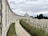 An expansive war memorial site with a long, curving wall of engraved names. A central domed structure stands prominently in the distance, surrounded by neatly arranged rows of white gravestones. The foreground features a path bordered by vibrant red flowers and manicured grass, framed by tall, lush trees under a cloudy sky.
