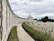 An expansive war memorial site with a long, curving wall of engraved names. A central domed structure stands prominently in the distance, surrounded by neatly arranged rows of white gravestones. The foreground features a path bordered by vibrant red flowers and manicured grass, framed by tall, lush trees under a cloudy sky.