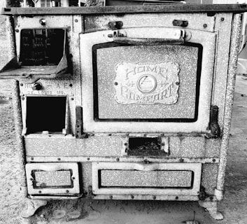 An antique cast iron stove labeled 'Home Comfort' with intricate detailing and multiple compartments. The stove appears weathered and is situated outdoors, emphasizing its historical and vintage aspect.