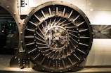 A large, round, metallic vault door with intricate spokes and a central locking mechanism dominates the image. The surroundings are industrial and metallic, featuring bolts and structured patterns.