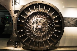 A large, round, metallic vault door with intricate spokes and a central locking mechanism dominates the image. The surroundings are industrial and metallic, featuring bolts and structured patterns.