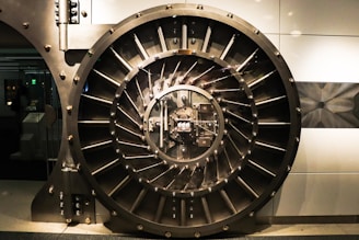 A large, round, metallic vault door with intricate spokes and a central locking mechanism dominates the image. The surroundings are industrial and metallic, featuring bolts and structured patterns.