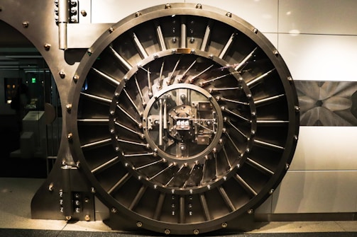A large, round, metallic vault door with intricate spokes and a central locking mechanism dominates the image. The surroundings are industrial and metallic, featuring bolts and structured patterns.
