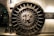 A large, round, metallic vault door with intricate spokes and a central locking mechanism dominates the image. The surroundings are industrial and metallic, featuring bolts and structured patterns.