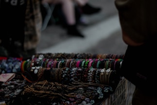 A collection of handcrafted bracelets displayed on a table, featuring a variety of colors, patterns, and materials such as beads and shells. The background is dimly lit, suggesting an outdoor market or street setting. The bracelets are neatly arranged in rows, demonstrating a diverse range of styles.