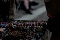 A collection of handcrafted bracelets displayed on a table, featuring a variety of colors, patterns, and materials such as beads and shells. The background is dimly lit, suggesting an outdoor market or street setting. The bracelets are neatly arranged in rows, demonstrating a diverse range of styles.