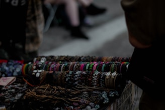 A collection of handcrafted bracelets displayed on a table, featuring a variety of colors, patterns, and materials such as beads and shells. The background is dimly lit, suggesting an outdoor market or street setting. The bracelets are neatly arranged in rows, demonstrating a diverse range of styles.