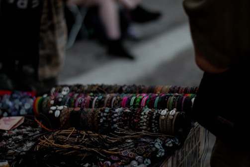 A collection of handcrafted bracelets displayed on a table, featuring a variety of colors, patterns, and materials such as beads and shells. The background is dimly lit, suggesting an outdoor market or street setting. The bracelets are neatly arranged in rows, demonstrating a diverse range of styles.