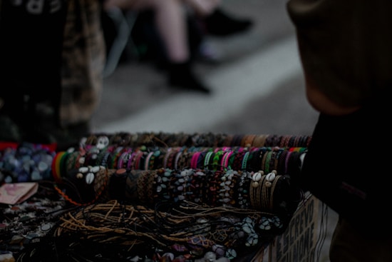 A collection of handcrafted bracelets displayed on a table, featuring a variety of colors, patterns, and materials such as beads and shells. The background is dimly lit, suggesting an outdoor market or street setting. The bracelets are neatly arranged in rows, demonstrating a diverse range of styles.