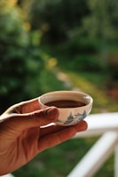A hand holds a small porcelain cup of tea with intricate blue designs against a backdrop of lush green foliage. Sunlight creates a warm atmosphere, highlighting the delicate craftsmanship of the cup.