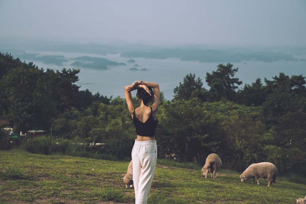 Frau, die in der Nähe von Schafen auf grünem Feld steht, umgeben von hohen und grünen Bäumen