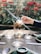 A hand holds a glass teapot with herbal tea inside, placed on a glass burner stand. The teapot is surrounded by three empty glass cups on saucers on a wooden table. In the background, there are red flowers and green foliage, creating a serene outdoor setting.