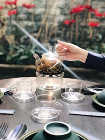 A hand holds a glass teapot with herbal tea inside, placed on a glass burner stand. The teapot is surrounded by three empty glass cups on saucers on a wooden table. In the background, there are red flowers and green foliage, creating a serene outdoor setting.