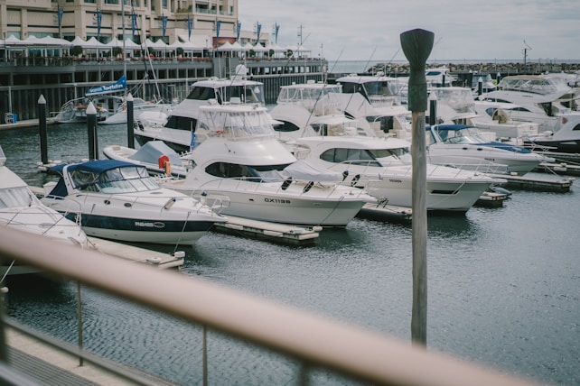 Multiple luxury yachts are docked in a marina, surrounded by calm water. A building with several balconies and blue flags is visible in the background, adding to the coastal setting. The scene is peaceful, with an overcast sky and various boats lined up neatly along the piers.