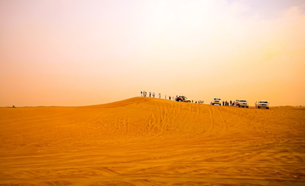 A vast desert landscape with golden sand dunes stretching into the distance. Several off-road vehicles are parked on one of the dunes with people standing around them, likely enjoying a desert excursion.