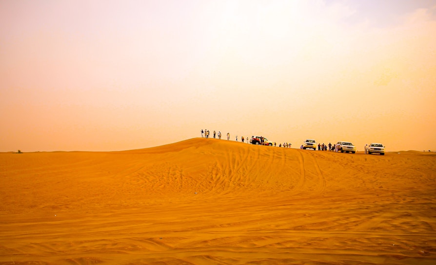 A vast desert landscape with golden sand dunes stretching into the distance. Several off-road vehicles are parked on one of the dunes with people standing around them, likely enjoying a desert excursion.