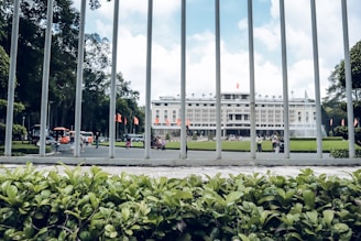 A large government or historical building in the background surrounded by a well-maintained garden. In the foreground, metal bars from a fence and lush green plants are visible. Several people are walking near the building, and the sky is partly cloudy.