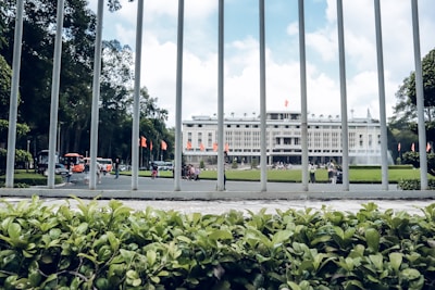 A large government or historical building in the background surrounded by a well-maintained garden. In the foreground, metal bars from a fence and lush green plants are visible. Several people are walking near the building, and the sky is partly cloudy.