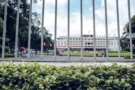 A large government or historical building in the background surrounded by a well-maintained garden. In the foreground, metal bars from a fence and lush green plants are visible. Several people are walking near the building, and the sky is partly cloudy.