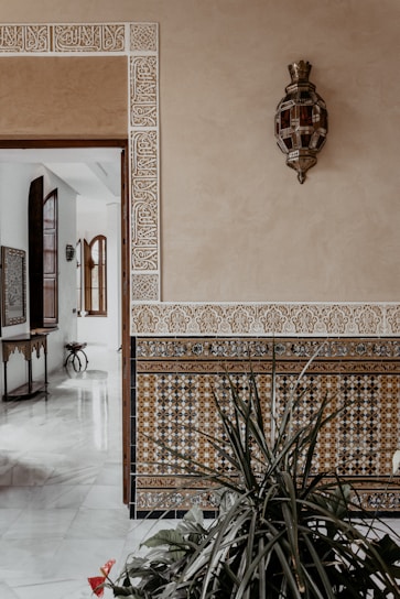 A beautifully decorated interior features a beige wall with intricate geometric tile patterns along the lower half and ornate white engravings around the doorway. There is a decorative wall lamp with an intricate design. In the foreground, lush green plants add a natural touch to the setting.