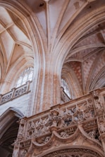A highly detailed interior of a historical building showcasing intricate architectural features. The scene includes tall, rib-vaulted ceilings, ornate stone arches, and elaborate carvings. Large, arched windows allow light to filter through, illuminating the stone structure and highlighting the fine craftsmanship of the design.