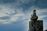 A small statue of a meditative figure sitting on a wooden post, surrounded by a vast, cloudy sky. The figure appears serene and contemplative, facing into the open expanse.