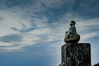 A small statue of a meditative figure sitting on a wooden post, surrounded by a vast, cloudy sky. The figure appears serene and contemplative, facing into the open expanse.