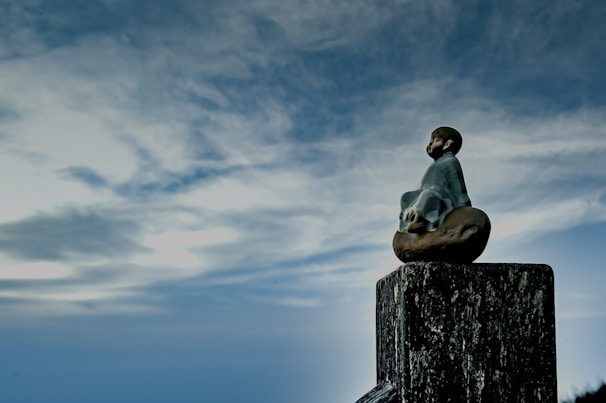 A small statue of a meditative figure sitting on a wooden post, surrounded by a vast, cloudy sky. The figure appears serene and contemplative, facing into the open expanse.