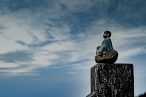 A small statue of a meditative figure sitting on a wooden post, surrounded by a vast, cloudy sky. The figure appears serene and contemplative, facing into the open expanse.