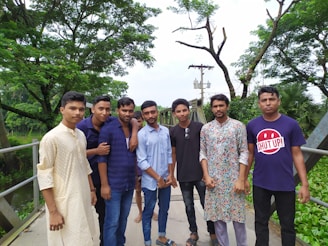 A group of young men standing on a metal bridge surrounded by lush green trees. They are wearing a mix of traditional and casual clothing. The background shows greenery, trees, and some power lines.