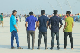Five young men stand in a row, facing away, on a busy promenade. The setting appears to be an outdoor public space with people walking and colorful umbrellas in the background. The men are wearing casual clothes in various shades, from blue and green tops to black and blue jeans.