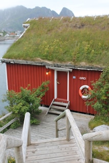 A quaint red wooden cabin is situated by the water, featuring a characteristic green grass roof. A wooden pathway with railings leads down to the entrance. A bright lifebuoy hangs on the side of the cabin, providing a vibrant contrast to the natural surroundings. Lush greenery surrounds the cabin, and mountains can be seen in the distance.