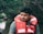 A man wearing a red life jacket is sitting in a natural, wooded area with water in the background. The background appears lush and green, suggesting a river or lake setting.