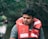 A man wearing a red life jacket is sitting in a natural, wooded area with water in the background. The background appears lush and green, suggesting a river or lake setting.
