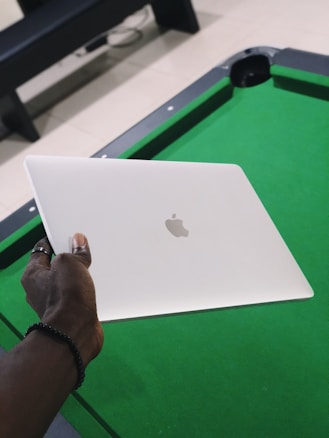 A hand holds a closed silver laptop with an Apple logo, positioned over a green pool table. The background includes a beige floor and part of a black bench.