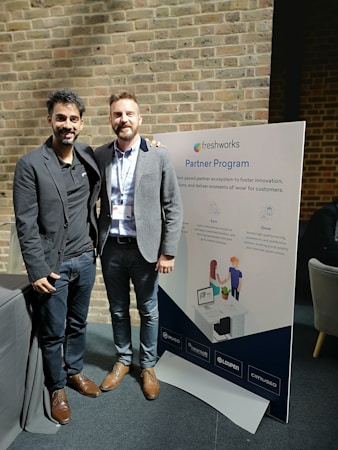 Two men are standing side by side, smiling, with one placing his arm around the other's shoulder. They are in a professional setting, dressed in business casual attire. Behind them is a sign that reads 'Freshworks Partner Program' with some description and logos. The background consists of a brick wall.
