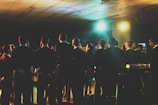 A group of people, dressed in formal attire, stand together, possibly participating in a choir or rehearsal. They are facing forward, away from the camera, and are illuminated by colorful stage lights. The setting appears to be indoors, in a large room with a tiled ceiling.