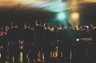A group of people, dressed in formal attire, stand together, possibly participating in a choir or rehearsal. They are facing forward, away from the camera, and are illuminated by colorful stage lights. The setting appears to be indoors, in a large room with a tiled ceiling.