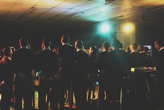 A group of people, dressed in formal attire, stand together, possibly participating in a choir or rehearsal. They are facing forward, away from the camera, and are illuminated by colorful stage lights. The setting appears to be indoors, in a large room with a tiled ceiling.