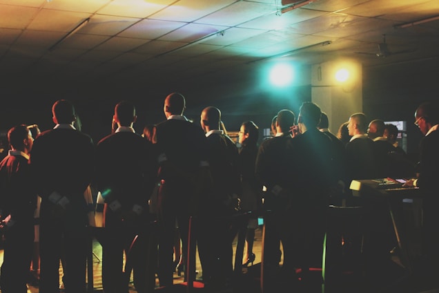 A group of people, dressed in formal attire, stand together, possibly participating in a choir or rehearsal. They are facing forward, away from the camera, and are illuminated by colorful stage lights. The setting appears to be indoors, in a large room with a tiled ceiling.