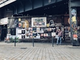 An urban street scene with a graffiti-covered wall displaying a variety of posters and street art beneath an overpass. A man stands beside the wall holding a drink, while a few bicycles are parked nearby. The atmosphere is gritty and artistic, reflecting a vibrant urban culture.