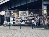 An urban street scene with a graffiti-covered wall displaying a variety of posters and street art beneath an overpass. A man stands beside the wall holding a drink, while a few bicycles are parked nearby. The atmosphere is gritty and artistic, reflecting a vibrant urban culture.