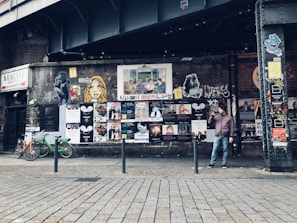An urban street scene with a graffiti-covered wall displaying a variety of posters and street art beneath an overpass. A man stands beside the wall holding a drink, while a few bicycles are parked nearby. The atmosphere is gritty and artistic, reflecting a vibrant urban culture.